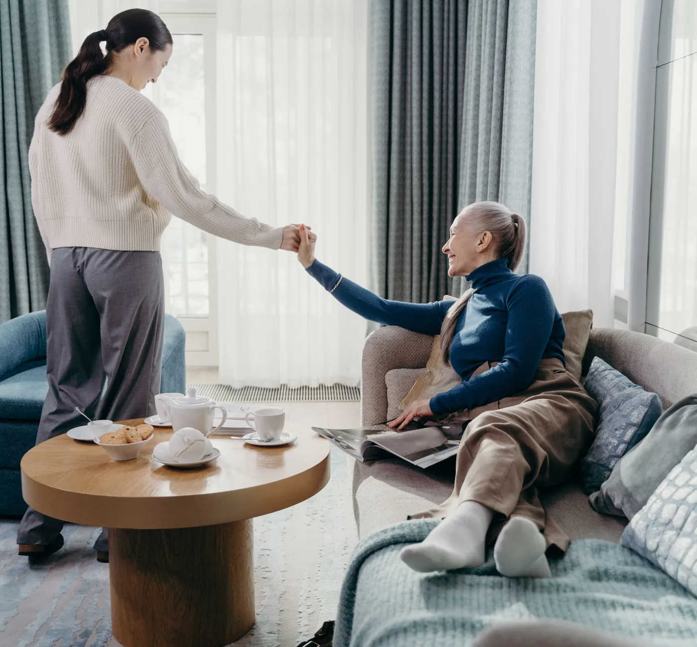 A woman helping her mother up from the sofa in a bright living room